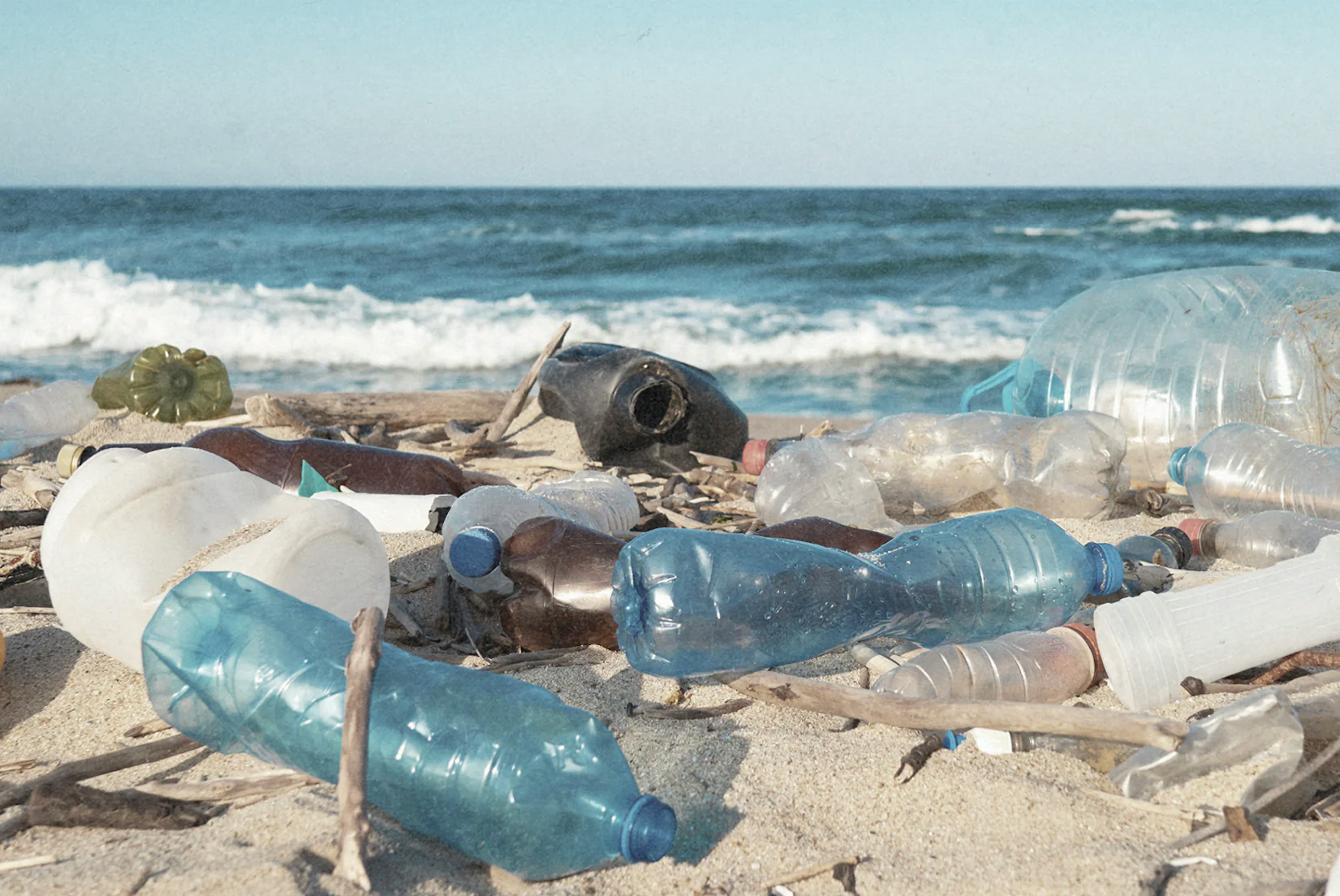 des déchets plastiques, surtout des bouteilles et bidons de différentes couleurs, sur une plage de sable blanc, proche des vagues et sous un ciel bleu, synonyme de la crise de pollution plastique.