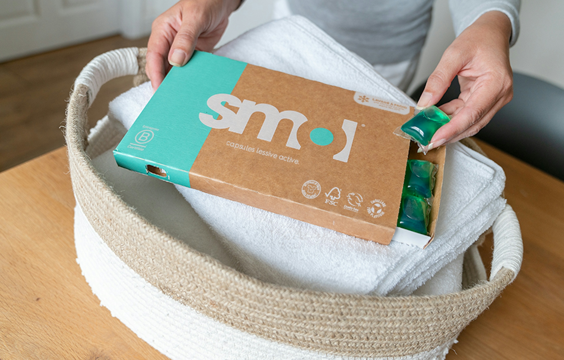 pack de capsules lessive active smol avec un tiroir ouvert, posé sur un panier de linge blanc propre, et une main tenant une capsule.