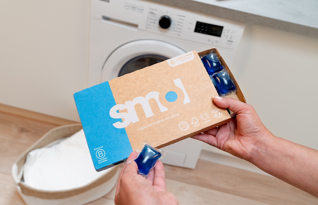 pack de capsules lessive sensitive smol tenue au-dessus d'un panier de linge blanc et d'une machine à laver.
