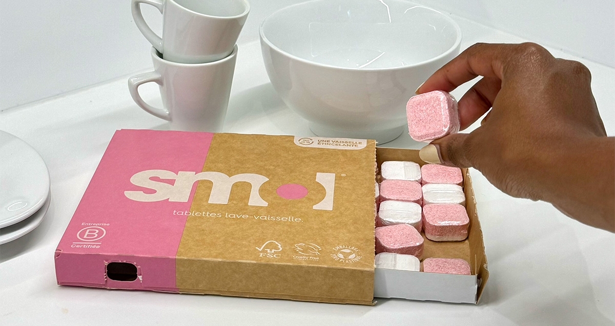 main prenant une tablette lave-vaisselle smol dans un pack au tiroir ouvert, posé sur une surface blanche à côté de vaisselle blanche.