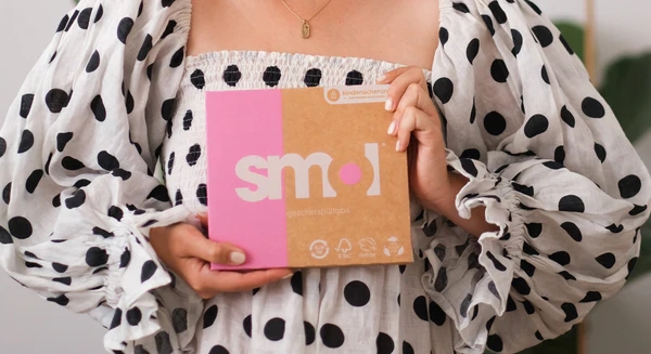 eine weiße Person in einem weißen Kleid mit schwarzen Punkten hält eine Packung smol Geschirrspültabs vor ihrem Körper