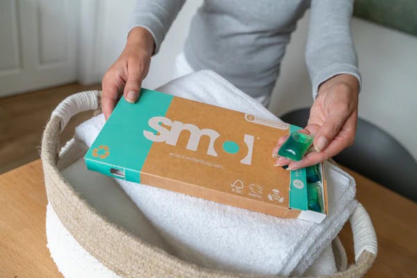 die Hände einer weißen Person, die eine halb offene Packung smol Universal Waschmittelkapseln über einem gefüllten Wäschekorb halten