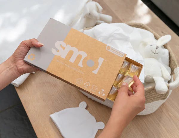 die Hände einer weißen Person, die eine halb offene Packung smol Sensitiv Waschmittelkapseln über einem gefüllten Wäschekorb halten