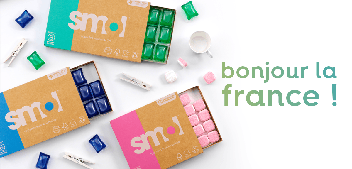 packs de capsules lessive active, de capsules lessive sensitive et de tablettes lave-vaisselle dont le tiroir est ouvert, révélant quelques capsules et tablettes. Des pinces à linge blanches et capsules sont réparties autour de pack. Le texte "bonjour la France !" est sur la droite.