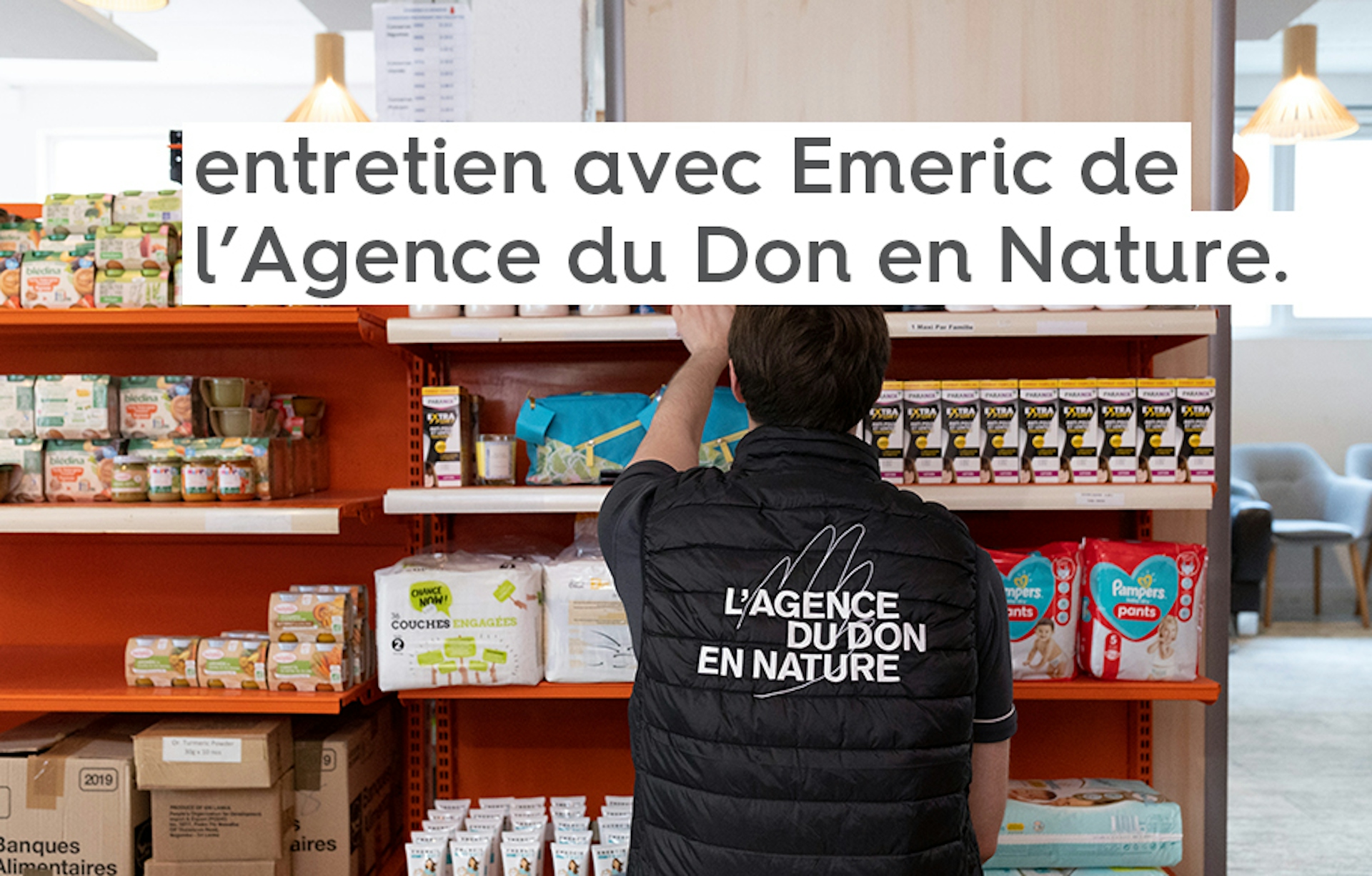 Un volontaire pour l’Agence du Don en Nature de dos portant un manteau bleu marine avec le logo de l’association, déposant des produits sur une étagère de magasin solidaire.