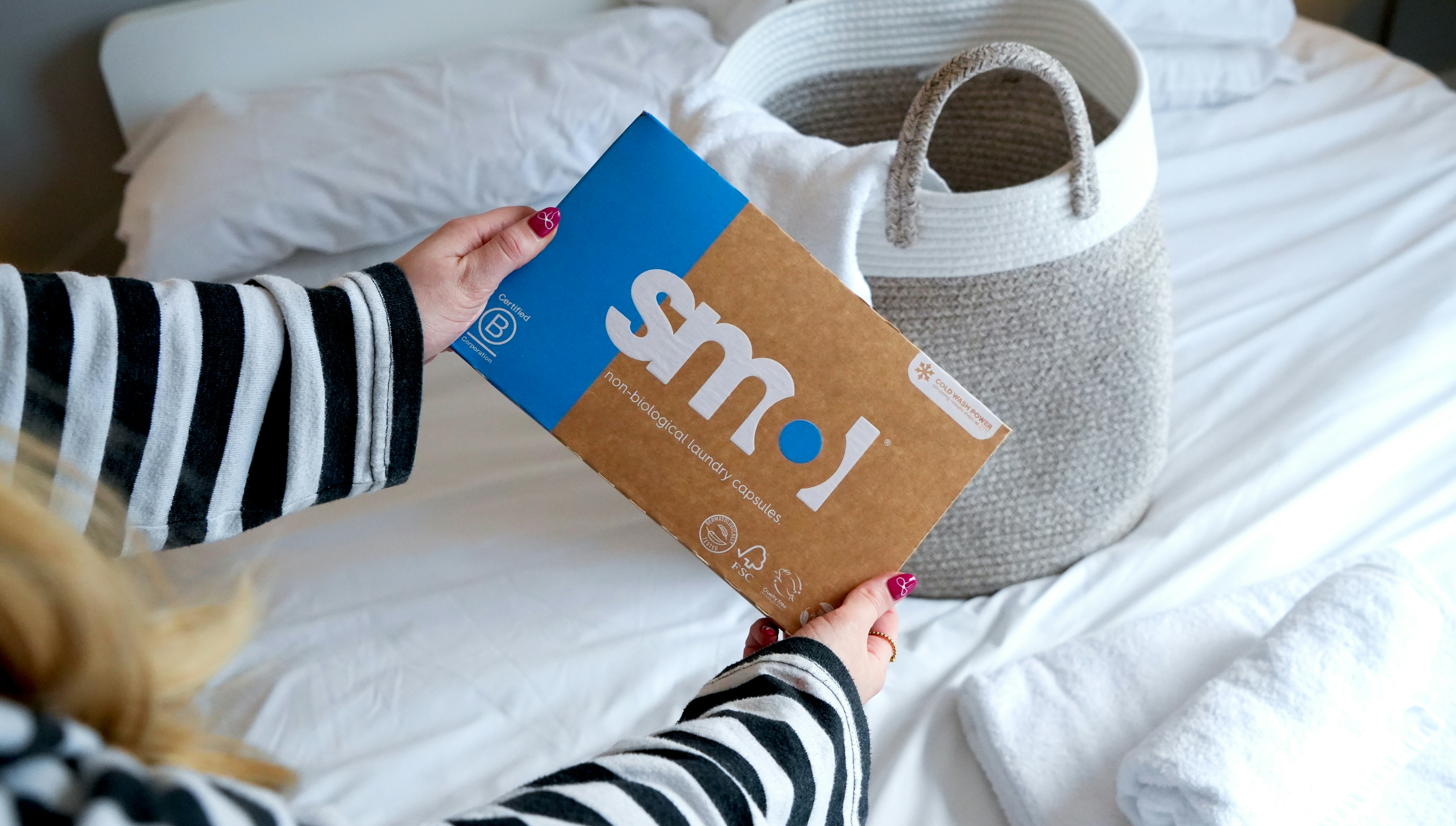 smol non bio laundry capsules held up in front of a bed with white sheets and a grey laundry basket