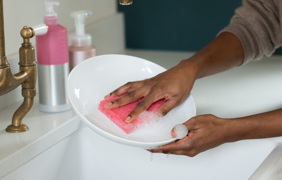 hands washing up a white plate in a sink using a pink smol sponge