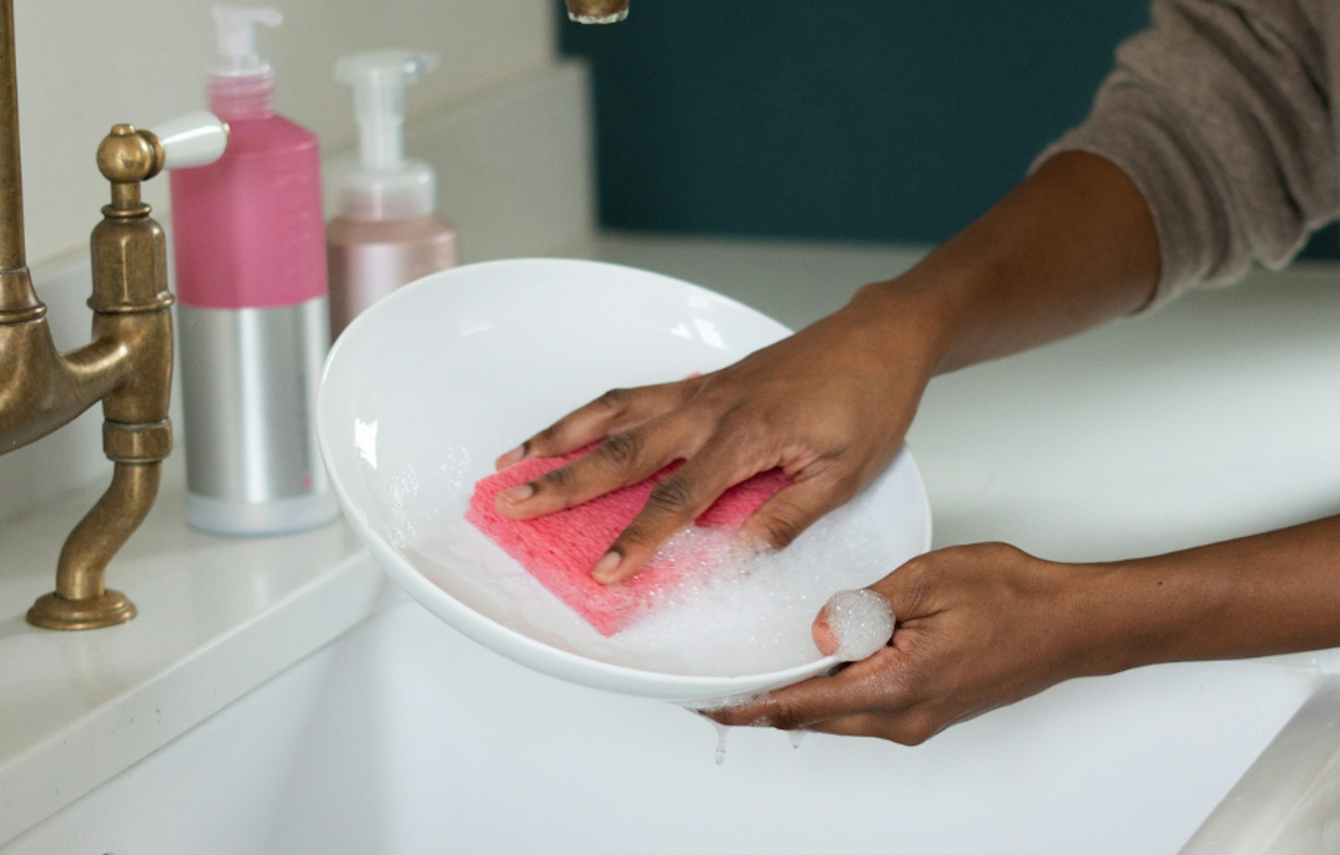 hands washing up a white plate in a sink using a pink smol sponge