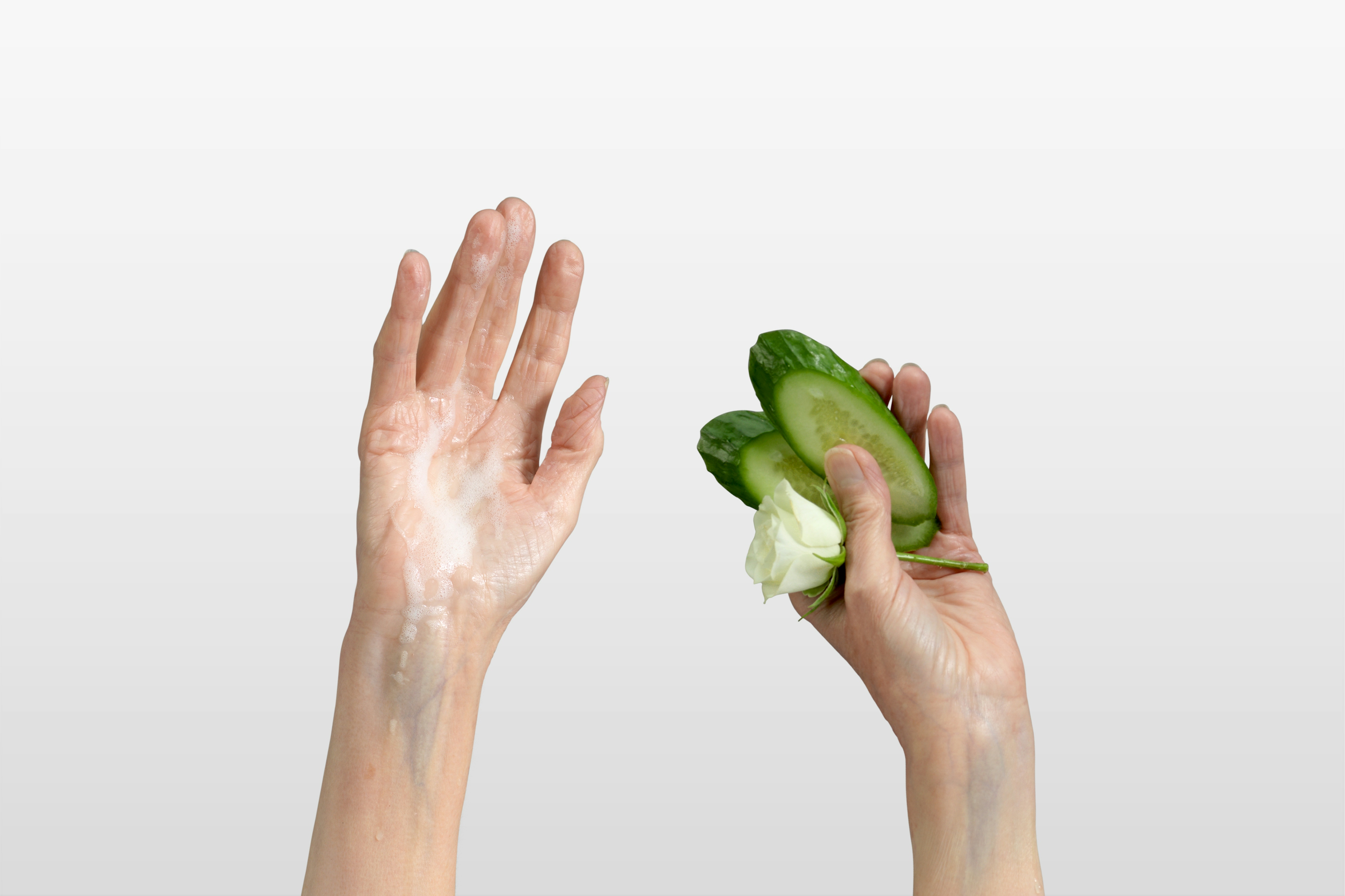 Set of hands up in the air, one hand covered in foam the other one holding cucumber slices and a rose