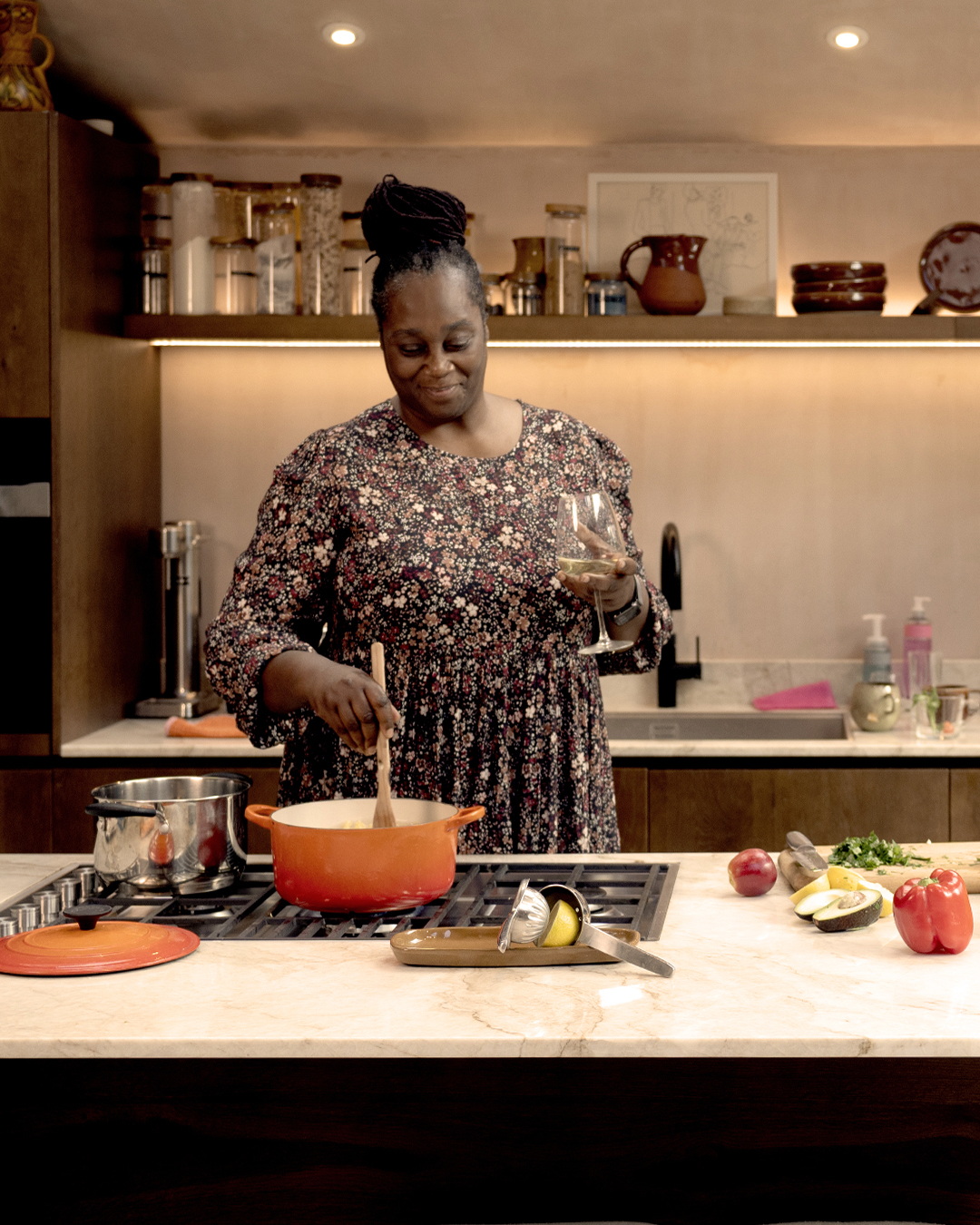 a smiling woman stirring a pot over a hob while holding a glass of wine