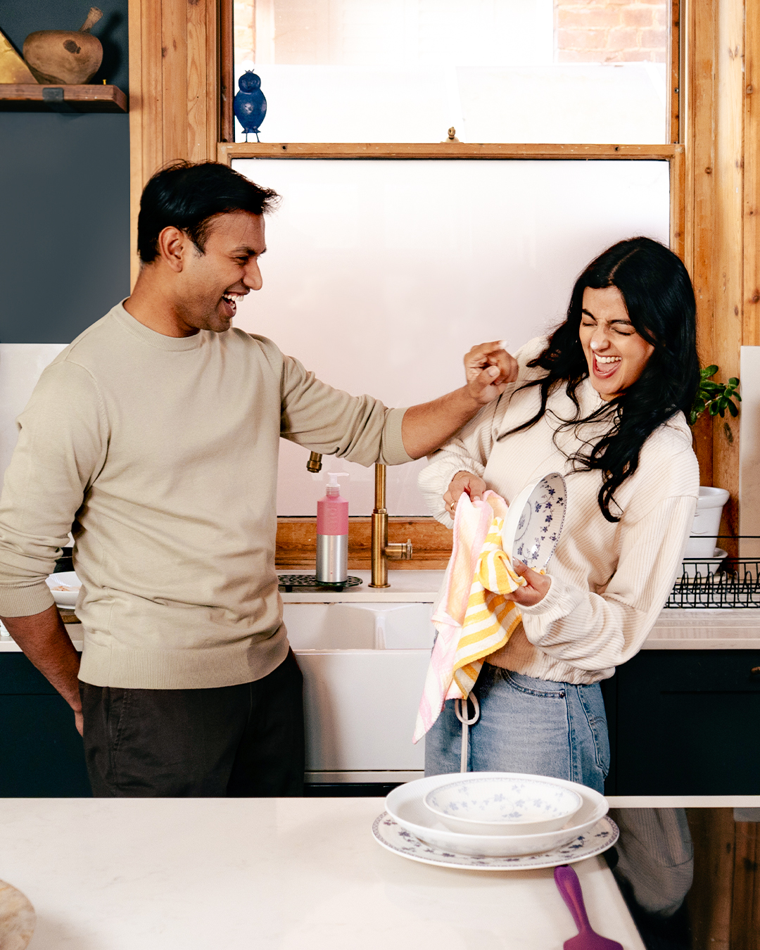 a couple standing by a kitchen sink laughing, the man is putting washing up soap bubbles on the woman's nose while she dries up the dishes