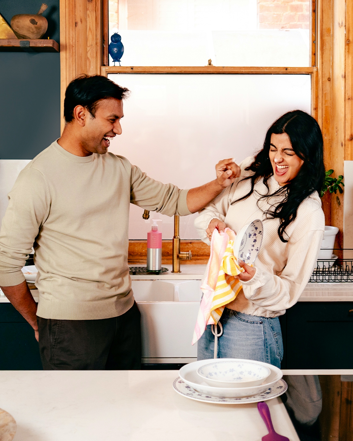 a couple standing by a kitchen sink laughing, the man is putting washing up soap bubbles on the woman's nose while she dries up the dishes