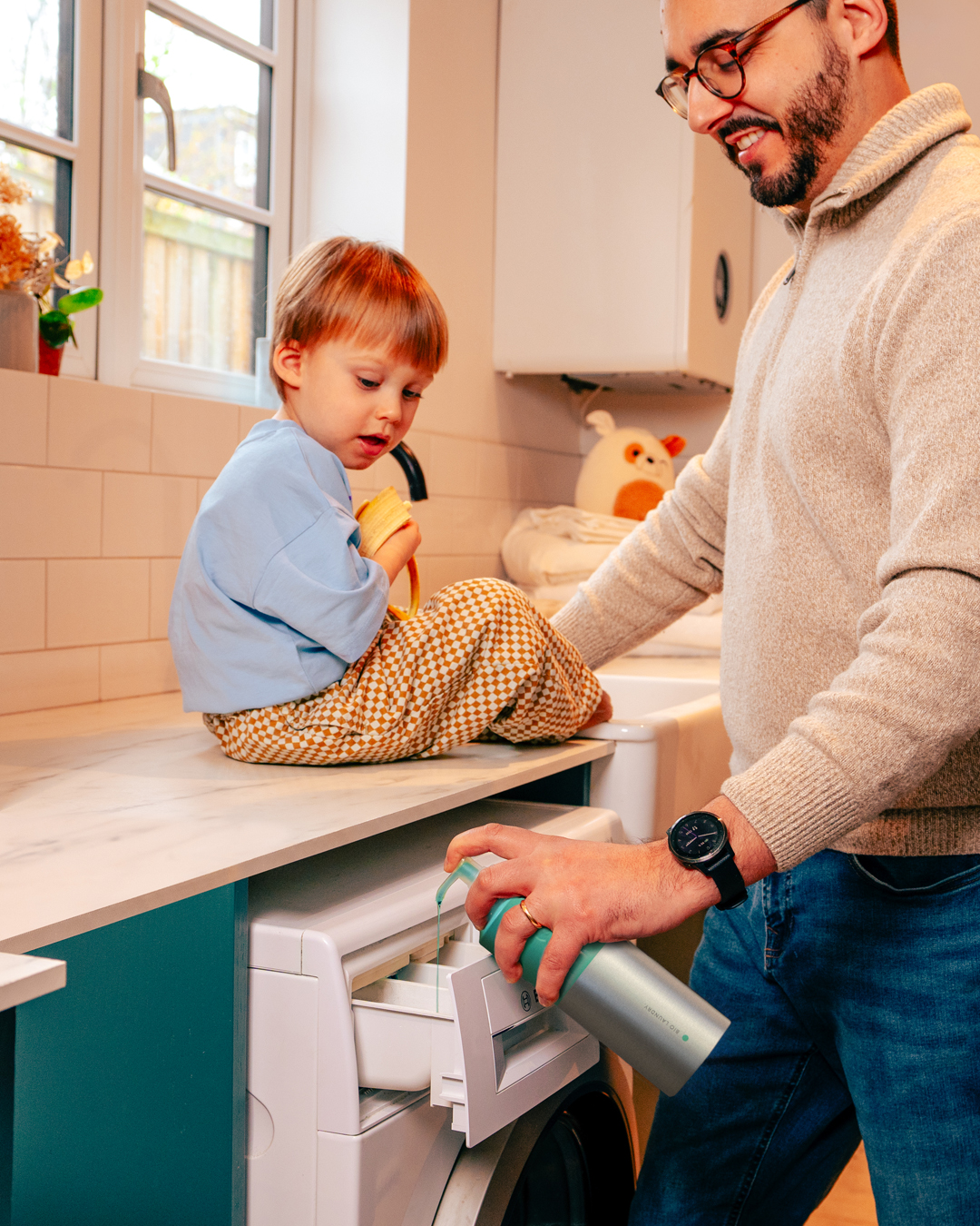A man pumping smol Laundry Liquid into his washing machine detergent drawer. His young child sits on the work surface next to him.