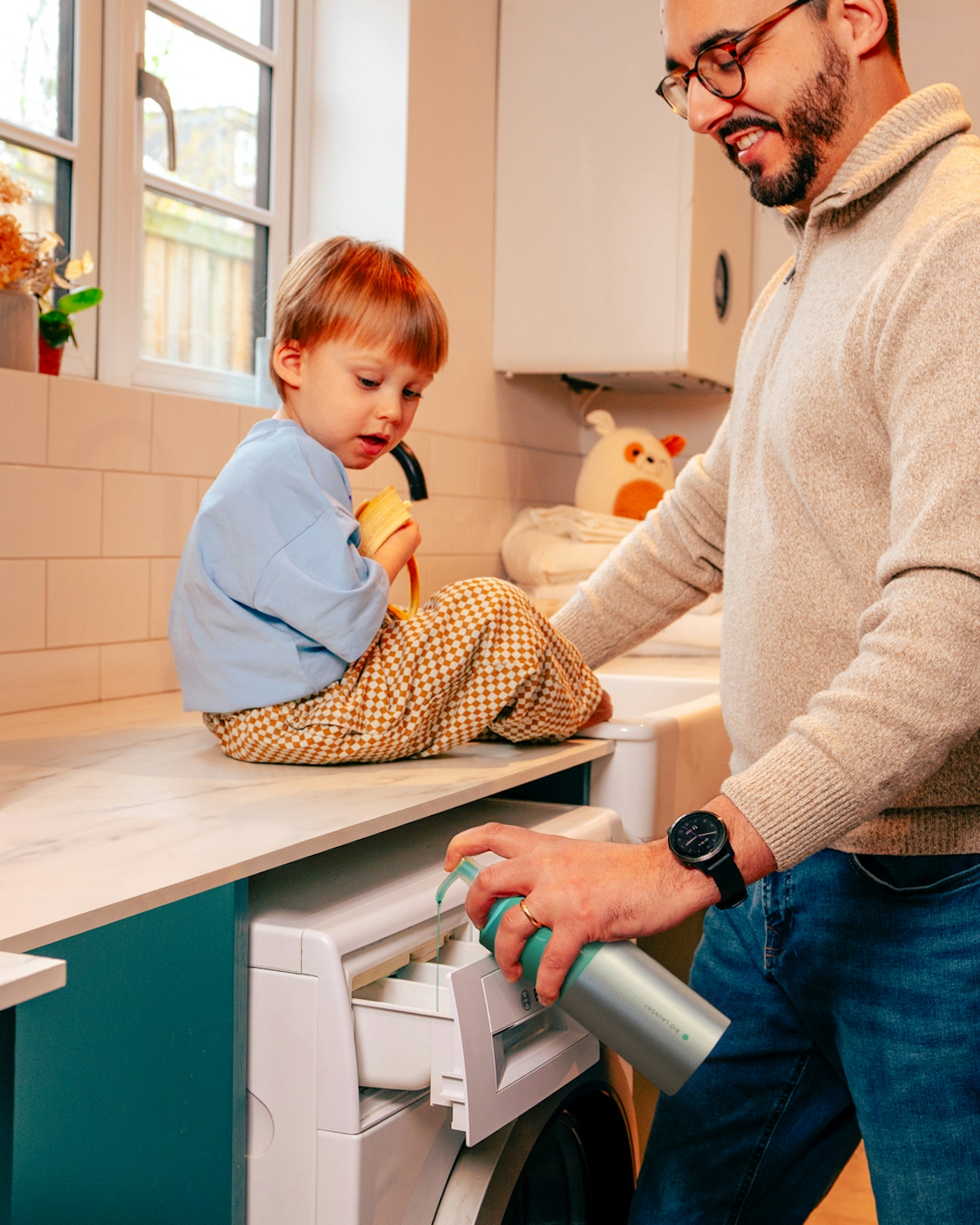 A man pumping smol Laundry Liquid into his washing machine detergent drawer. His young child sits on the work surface next to him.
